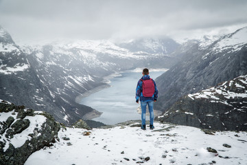 Man traveller with backpack standing back on edge and looking at scenery mountain landscape under snow, Trolltunga, Norway in may.