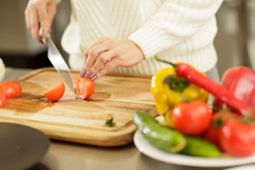 Cropped close up shot of a woman chopping tomatoes on the wooden cutting board, using sharp steel knife. Housewife making salad for her family, cooking at the kitchen