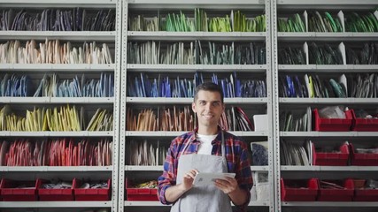 Artisan in checkered shirt smiling and looking at camera while standing near shelves with mosaic samples and using tablet - Powered by Adobe