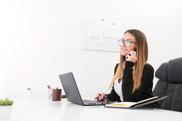 Portrait of young beautiful accountant talking on smartphone while browsing through some files in laptop at workplace.