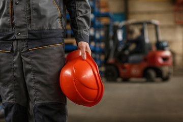 Cropped shot of a mid section of a male industrial worker wearing uniform holding protective hardhat, standing at the storage of metalworking company. Heavy industry concept