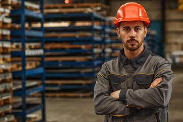 Horizontal portrait of a young male metalworker in protective hardhat and uniform posing confidently at the warehouse of a industrial manufacturing. Confidence, success, business concept