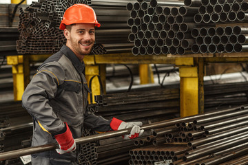 Horizontal portrait of a cheerful industrial worker in protective uniform and hardhat smiling to the camera over his shoulder while stacking steel pipes at the storage of a metalworking company