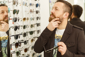 Portrait of a mature handsome bearded male customer rubbing his beard thoughtfully, while choosing...