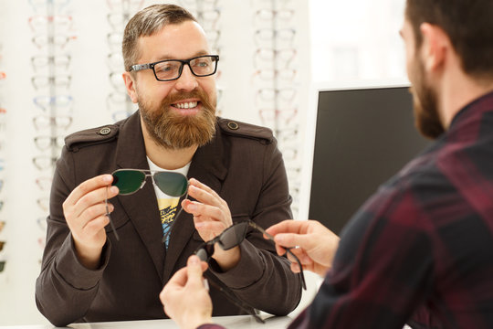 Cheerful matue man smiling joyfully, talking to the professional optometrist while choosing new sunglasses at the optician shop. Consumerism, communication, sales, service concept