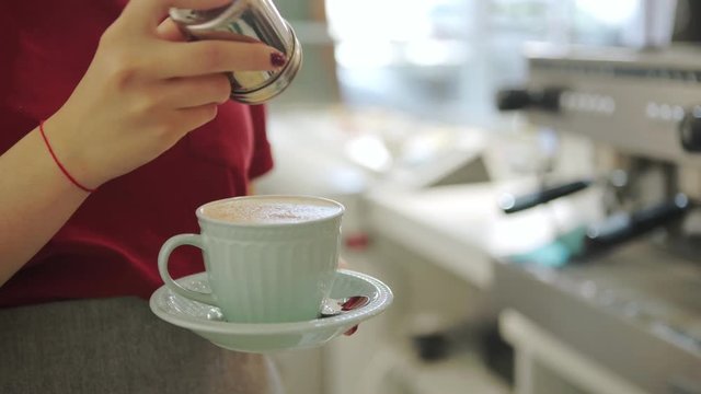 Anonymous female in uniform shaking spice dispenser over small cup of freshly brewed coffee while working in modern cafe.