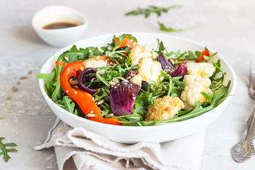 Salad with arugula, roasted vegetables (cauliflower, red bell pepper and onion) and seeds. Healthy food. Light grey concrete background. Copy space.