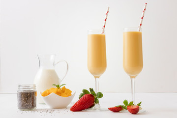 The porcelain bowl with pieces of fresh mango on the white wooden table and 2 glasses of fresh mango smoothie, or mango milkshake and jar of chia seeds and few real fresh strawberries.