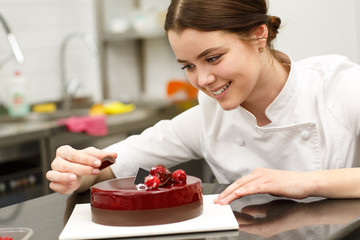Beautiful and cute girl smiling and decorating glazed cake. Young confectioner wearing white uniform while working in confectionery. She baking delicious dark red with chocolate. - obrazy, fototapety, plakaty