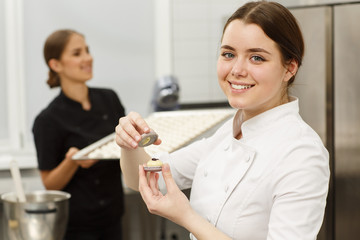 Happy and smiling female confectioner in white uniform holding delicious macarons in hands. Another confectionery in black uniform holding tray with macarons on background.