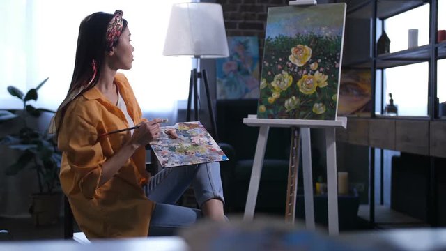 Side view of female painter holding palette and sitting near her finished artwork with yellow peonies on easel. Talented woman artist ponding the final touches of masterpiece in home art studio