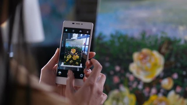 Close up hands of female painter holding cellphone photographing artwork on easel in home art studio. Artist hands holding mobile phone and making a picture o flowers painting on phone screen