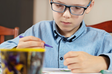 Skupiona twarz chłopca w niebieskiej koszuli i okularach podczas malowania obrazka. Chłopiec trzyma flamaster w dłoni.  