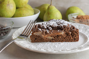 Close up side view of Chocolate Pear Cake sprinkled with powdered sugar on white plate.  Chocolate chips to the left, cocoa powder in back right corner, pears in white bowl in back  left corner.