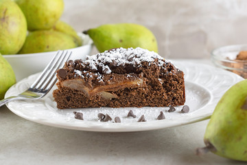 Close up side view of Chocolate Pear Cake sprinkled with powdered sugar on white plate.  Chocolate chips and fork on plate.  Pears and cocoa powder in background.