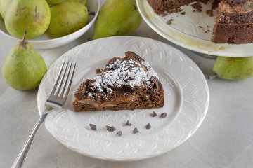 Top side view of Chocolate Pear Cake on a white plate sprinkled with powdered sugar and chocolate chips.  Pears and full cake in background.