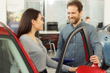Positivity and friendly man manager showing young happy woman customer cars. Beautiful smiling girl sitting inside vehicle in red car. Bearded sales consultant working in car dealership.