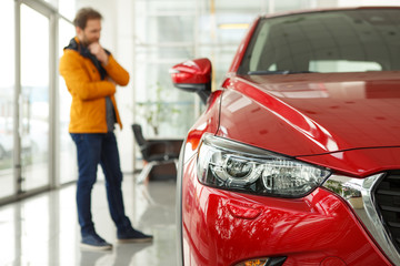Front view of headlight of beautiful red automobile. Male customer choosing cars and thinking about features on background. Concept of car dealership, purchase and service.