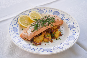 Salmon fillet or steak served with roasted potatoes and onion, slices of fresh lemon and chopped fresh dill on the rustic plate. Fresh, healthy, light, diet fish dish for lunch or diner.