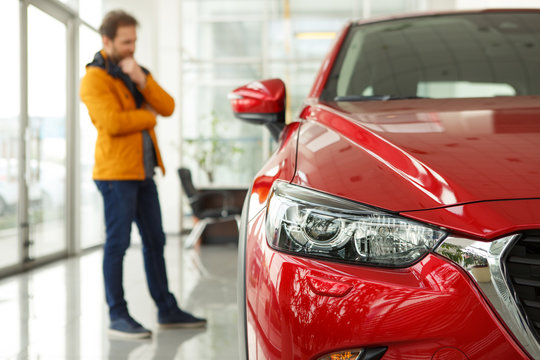 Front View Of Headlight Of Beautiful Red Automobile. Male Customer Choosing Cars And Thinking About Features On Background. Concept Of Car Dealership, Purchase And Service.