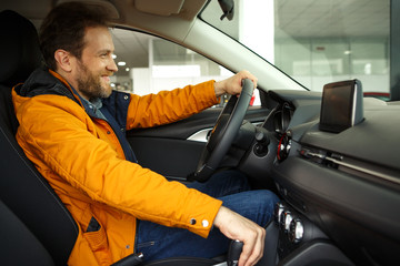 Male client sitting in automobile cabin and testing new car. Customer smiling and trying drive a car before purchase. Handsome man with beard wearing in yellow jacket.