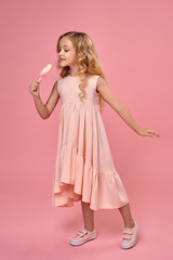 Fototapeta na wymiar Little girl with a blond curly hair, in a pink dress is posing with a candy