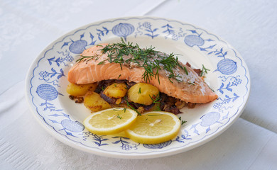 Salmon fillet or steak served with roasted potatoes and onion, slices of fresh lemon and chopped fresh dill on the rustic plate. Fresh, healthy, light, diet fish dish for lunch or diner.