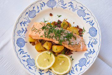 Top view on salmon fillet or steak served with roasted potatoes and onion, slices of fresh lemon and chopped fresh dill on the rustic plate. Fresh, healthy, light, diet fish dish for lunch or dinner.