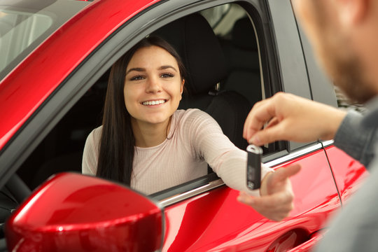Beautiful and young woman sitting in red car and smiling. Female customer holding her hand and receiving keys of her new car from manager who working in modern car dealership.