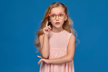 Fashionable little girl in a pink dress and glasses is standing on a blue background.