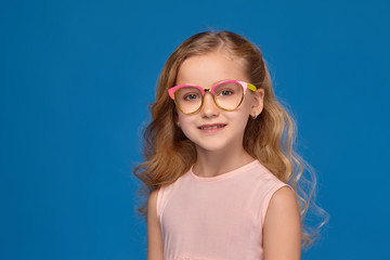 Fashionable little girl in a pink dress and glasses, with a candy in her hands, standing on a blue background.