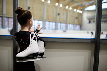 Back view portrait of little girl holding figure skates standing by ice rink and watching training,...