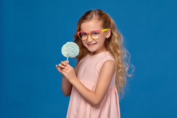 Fashionable little girl in a pink dress and glasses, with a candy in her hands, standing on a blue background.