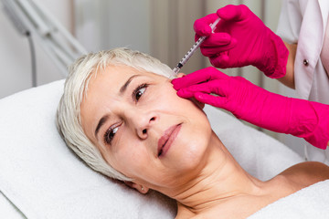 Obraz na płótnie Canvas Mature woman is getting a rejuvenating facial injections. She is lying calmly at clinic. The expert beautician is injecting botox into woman's wrinkles.