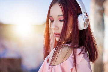 Gorgeous redheads lady listening music in headphones on blurred outdoor on background. Charming girl with white earphones enjoys with hair waving and eyes closed. Close up, selective focus