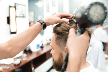 Side view of young bearded man getting groomed at hairdresser with hair dryer while sitting in chair at barbershop. The moment of styling hair dryer bearded man in the bright space of the barbershop