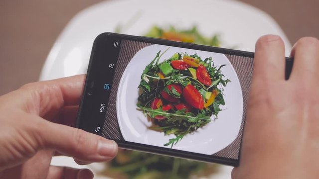 Female hands photographing a salad of ruccola, tomato and avocado with a smartphone.