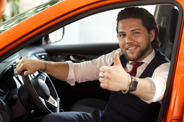 Customer of car dealership sitting in car, looking at camera and smiling. Bearded man showing thumb...