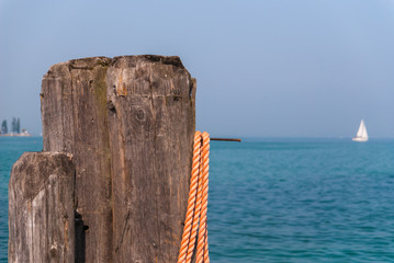 Wooden mooring pole with orange rope. In the background blurred Lake Garda with white sailboat