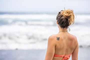 Cute girl portrait with long curly brown topknot hair and tattoo on the scapula while the woman is looking to the horizon at a wild beach with the crashing waves in the distance. Chile, South America