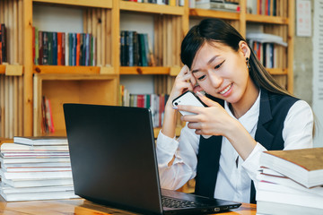 A cute Asian girl sits in the library, surrounded by books, working on a computer, reads a message on the phone. A teenager student prepares for exams, negotiates and learns.
