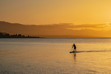 Naklejka na ściany i meble Silhouette Of A Man Paddleboarding Into Shore At Sunset
