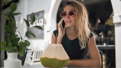 Fototapeta na wymiar Portrait of happy beautiful tanned female in cafe in open air, drink in a coconut on the table, girl chatting with her friends by modern phone, wearing fashionable sunglasses, copy space