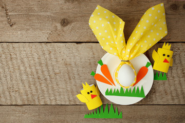 Easter children card concept with egg in bunny nupkin, white plate and paper craft diy decorative chikens on wooden background with copy space