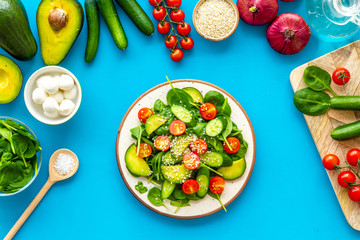 Ingredients for fresh salad. Vegetables, greens, spices, plate of salad on blue background top view