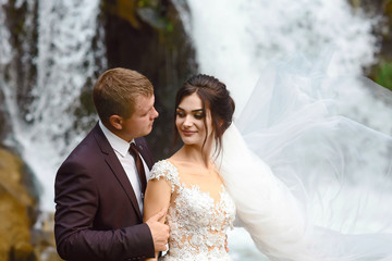 young wedding couple of sexy pretty girl or woman with hair in white bride dress and handsome man near beautiful grand waterfall in mountain.