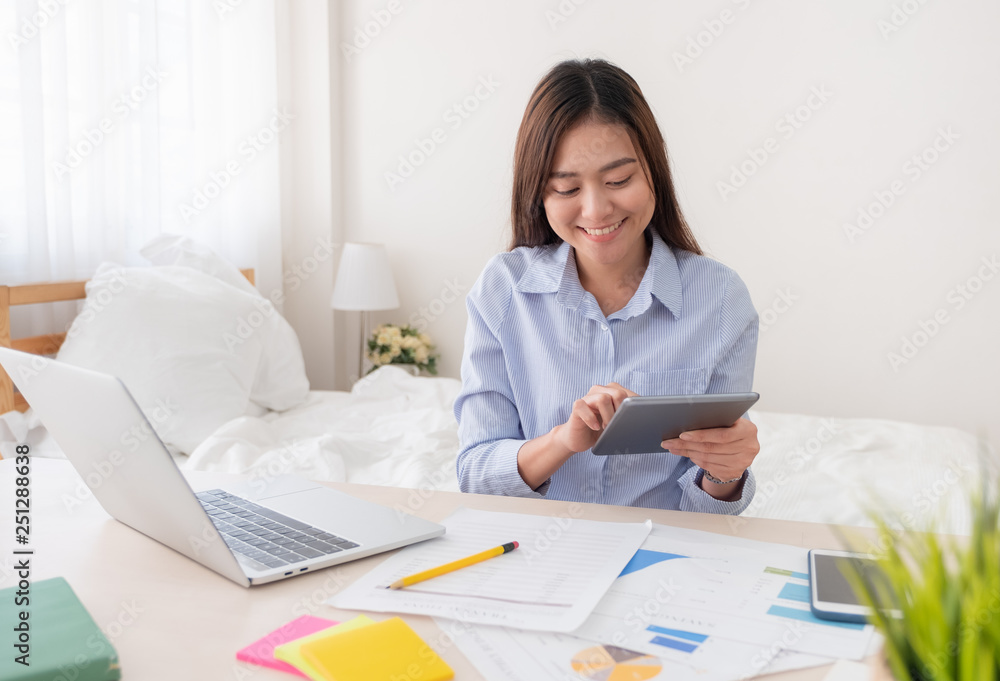 Poster Asian freelancer using tablet while working with laptop on table in bedroom at home.Work at home concept.work from home concept.