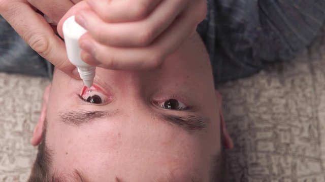 From above of adult man lying on couch and instilling drops into eyes and blinking
