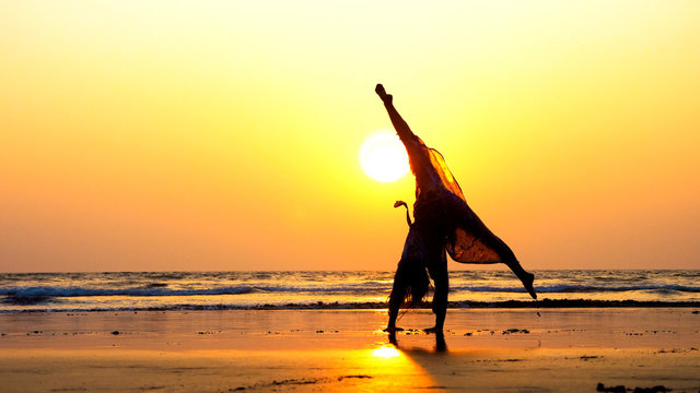 Silhouette of young gymnast woman doing handspring on sandy beach at beautiful orange sunset.