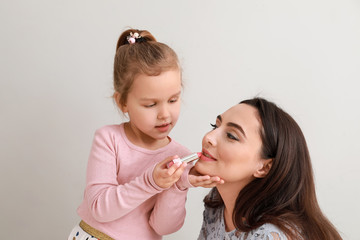 Cute little daughter applying lipstick onto mother's lips against light background